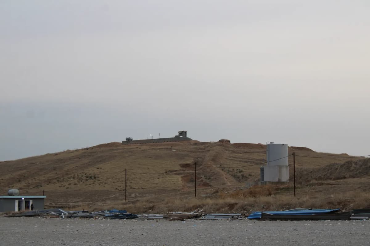 Colline aride surmontée d’un poste d’observation, réservoir blanc et embarcations échouées au premier plan.