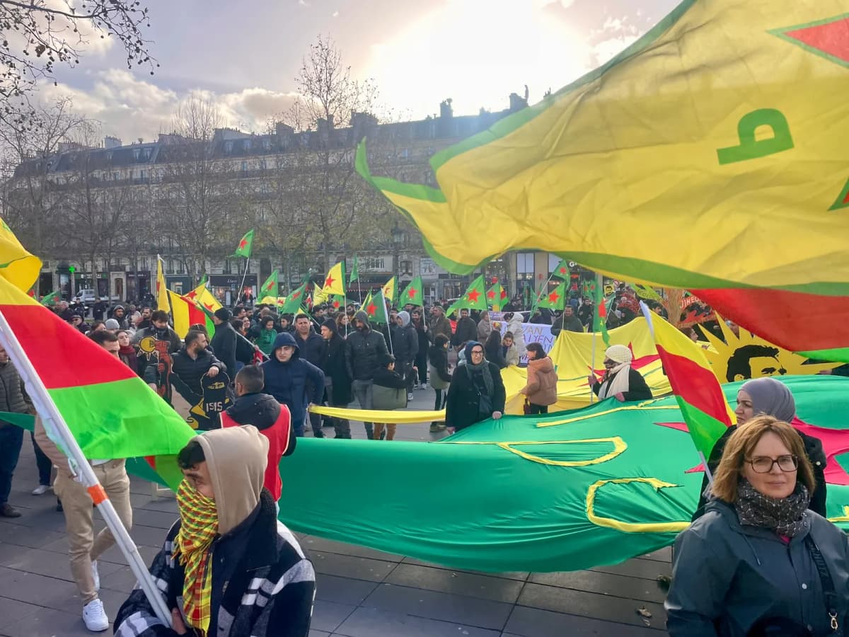 Manifestation sur une place, larges drapeaux verts, jaunes et rouges déployés au-dessus de la foule.