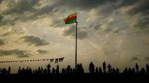 Drapeau vert, jaune et rouge flottant au sommet d’un mât, silhouettes d’une foule à contre-jour au crépuscule.