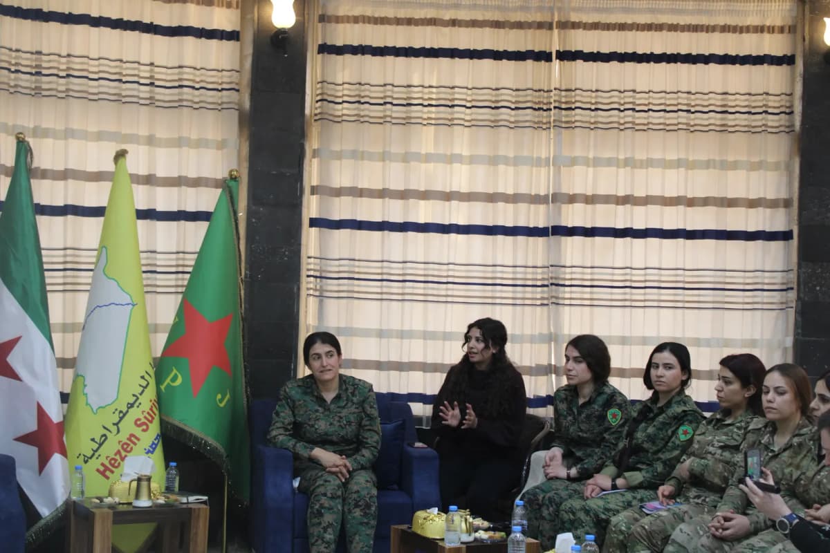 Plusieurs femmes en tenue militaire assises dans une salle, drapeaux et fanions posés à côté d’elles.