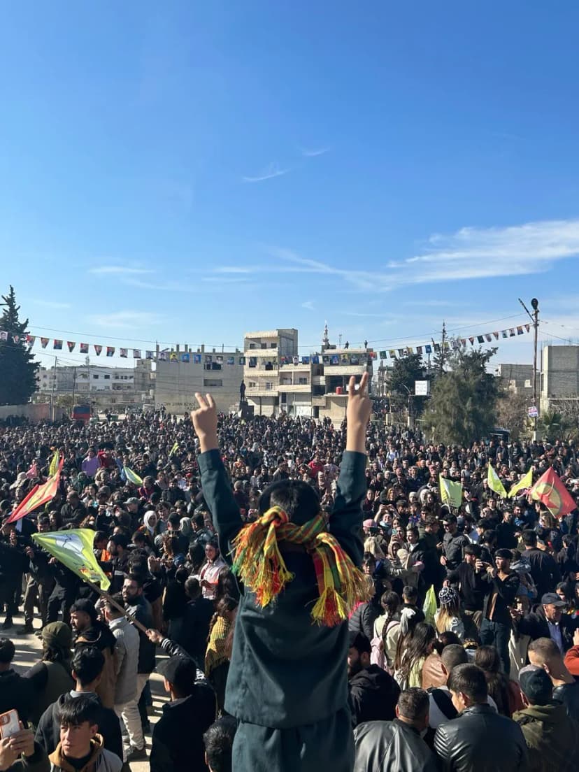 Foule dense rassemblée sur une place, une personne au premier plan levant les bras, drapeaux et guirlandes au-dessus.