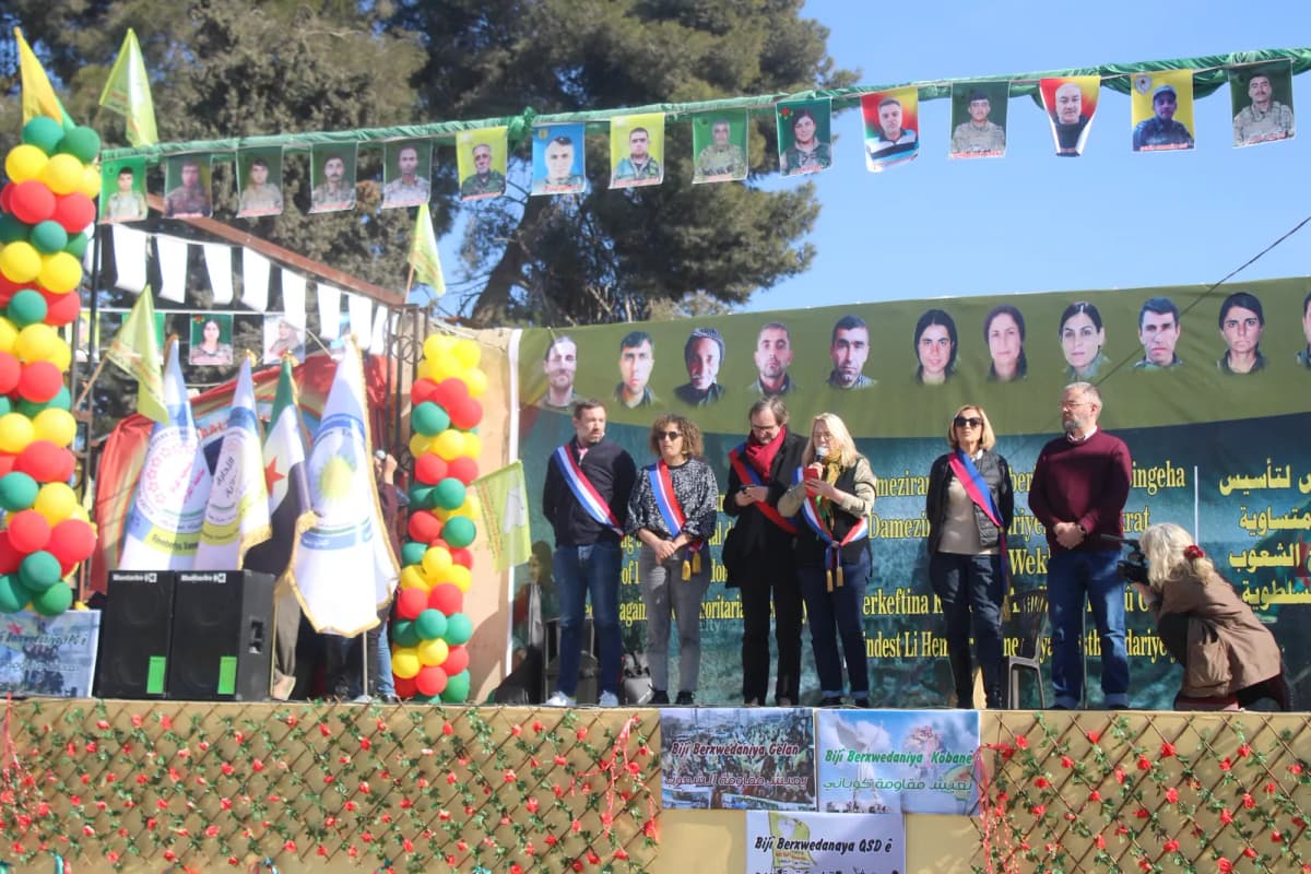 Estrade décorée de ballons devant une foule, plusieurs personnes en écharpe tricolore alignées sous un mur de portraits.