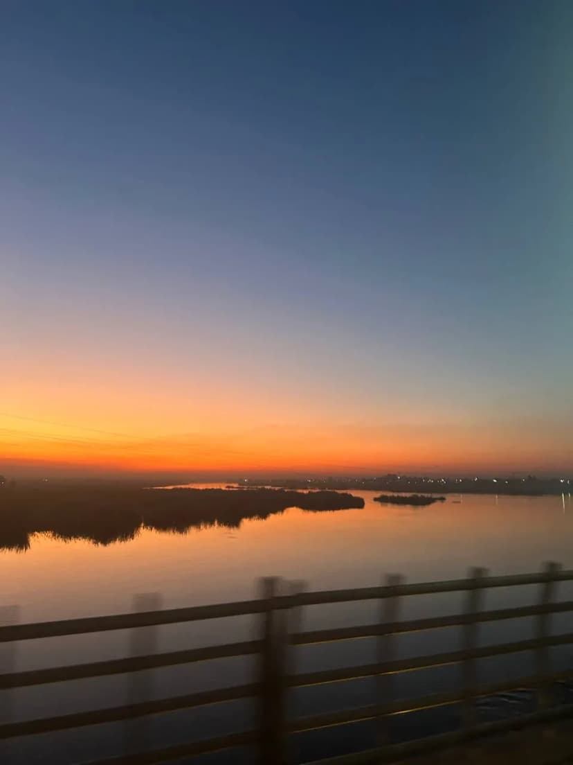 Coucher de soleil sur un fleuve, vu depuis un pont dont la rambarde barre le premier plan.