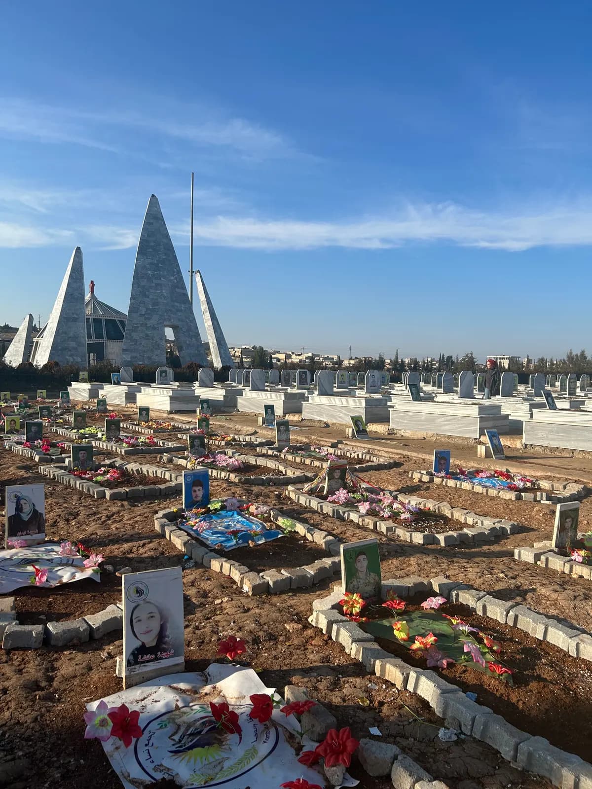 Cimetière aux tombes fleuries et photographies, monument aux flèches blanches à l’arrière-plan.