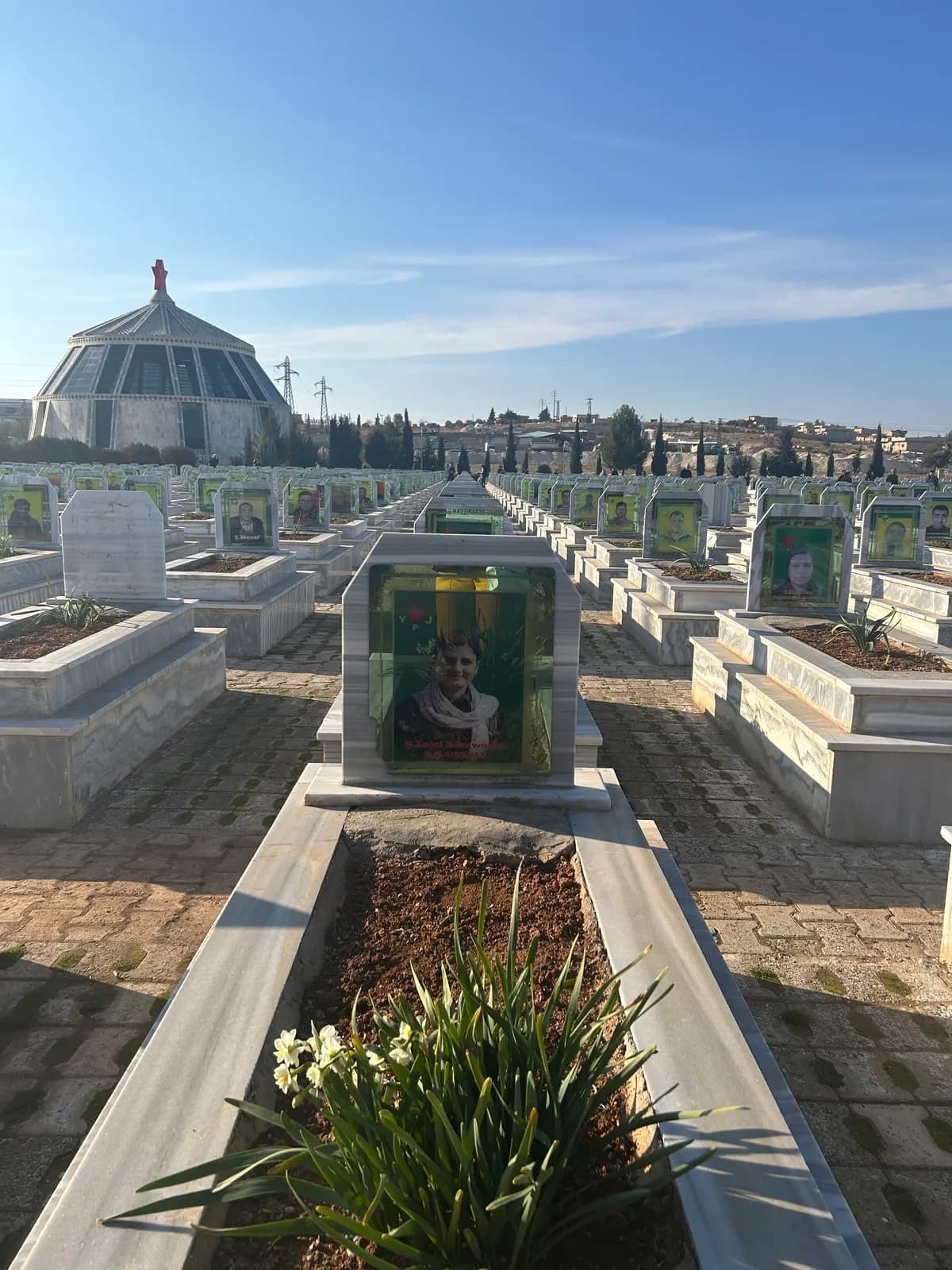 Allée d’un cimetière bordée de tombes portant des portraits, coupole d’un mémorial au fond.