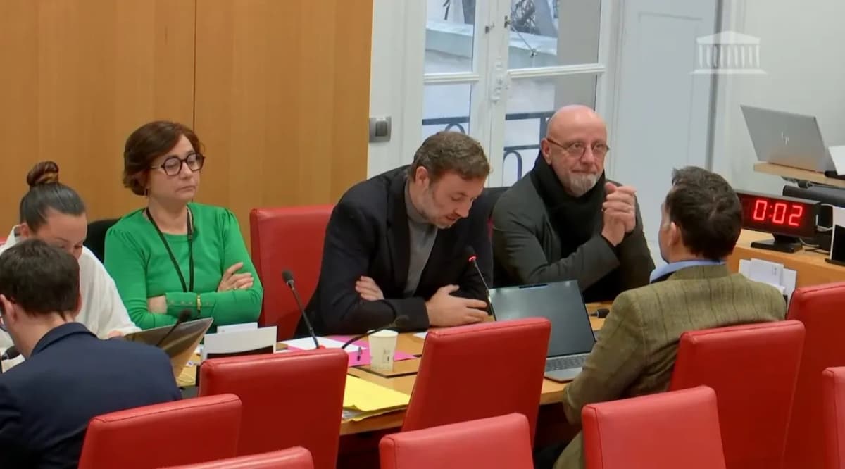 Réunion de commission à l’Assemblée nationale : des élus assis à une table aux fauteuils rouges, deux hommes se parlent.