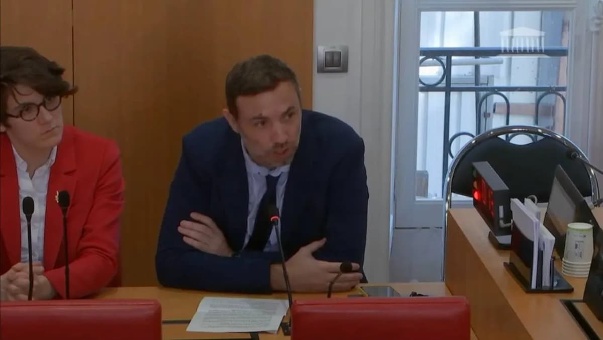 Réunion de commission : un homme bras croisés parle au micro à côté d’une femme en veste rouge.