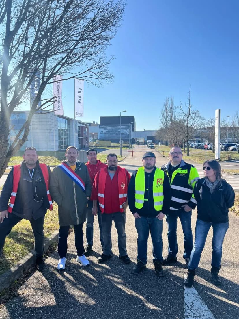 Sept personnes au bord d’une route devant un site logistique, plusieurs en chasuble syndicale, une en écharpe tricolore.