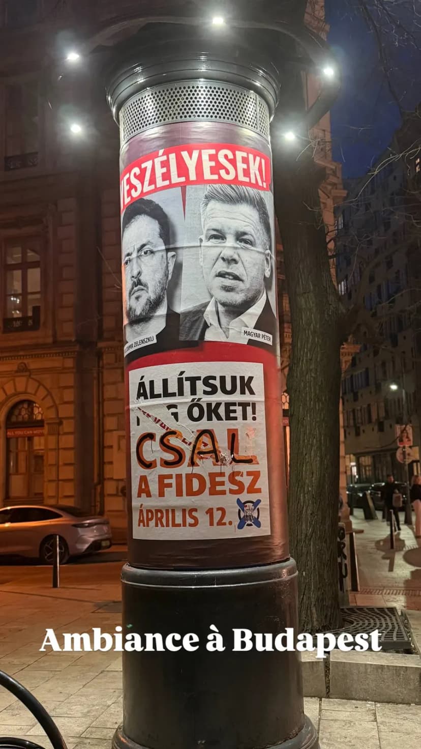 Colonne d’affichage éclairée dans une rue de nuit, couverte d’affiches politiques en hongrois.