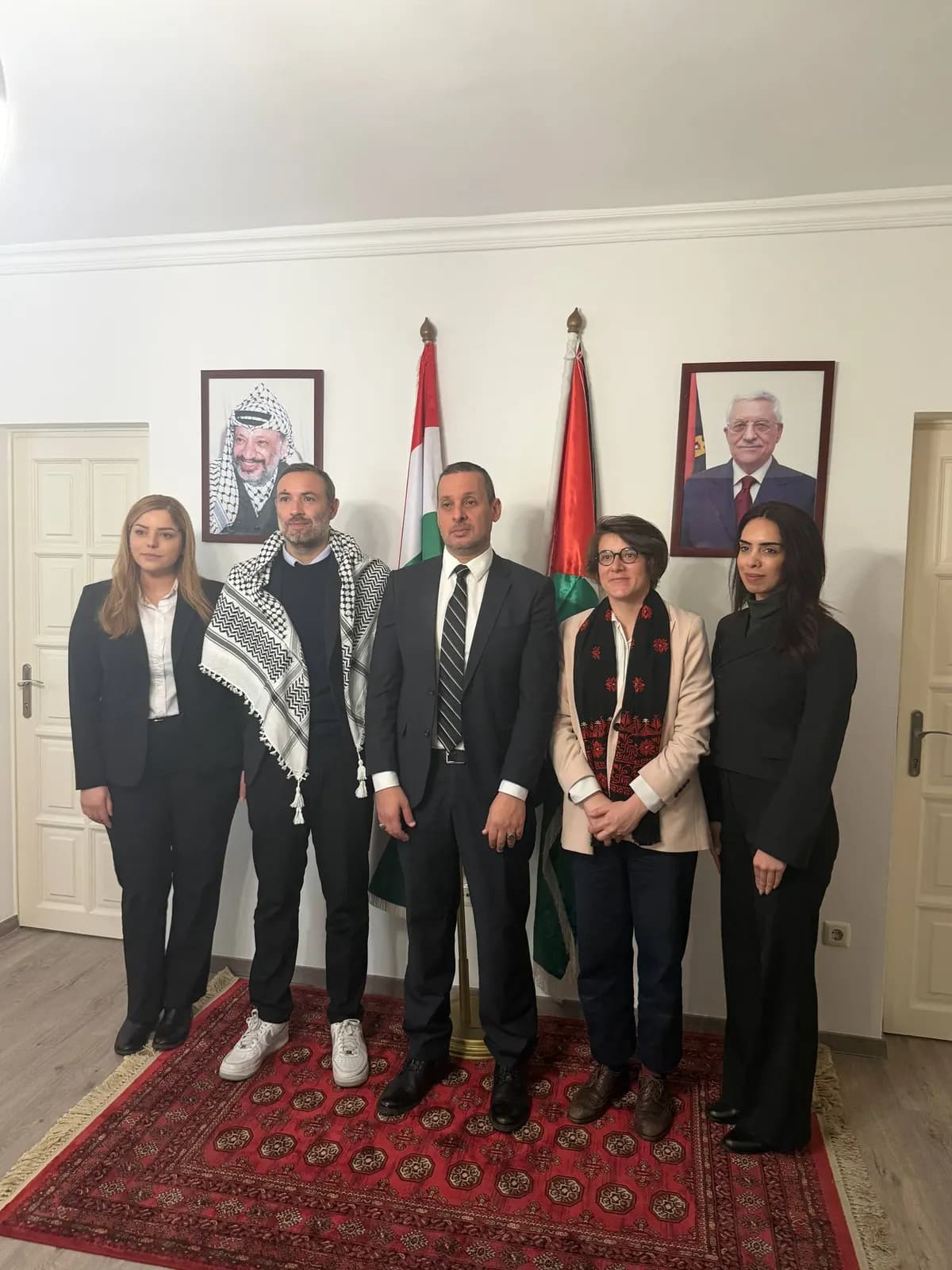Cinq personnes posent dans un salon d’ambassade, sous deux portraits officiels et entre deux drapeaux.