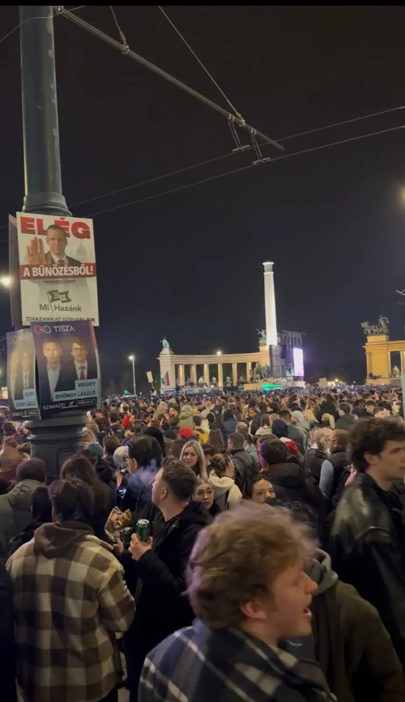 Foule dense rassemblée de nuit sur une grande place, colonnade éclairée à l’arrière-plan.
