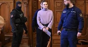 Une jeune femme en pull entourée d’agents en uniforme, l’un cagoulé, dans une salle boisée, le visage d’un agent flouté.