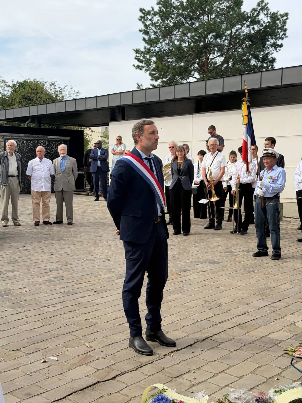 Un élu portant l’écharpe tricolore se tient devant un mémorial, face à des porte-drapeaux et à des participants.