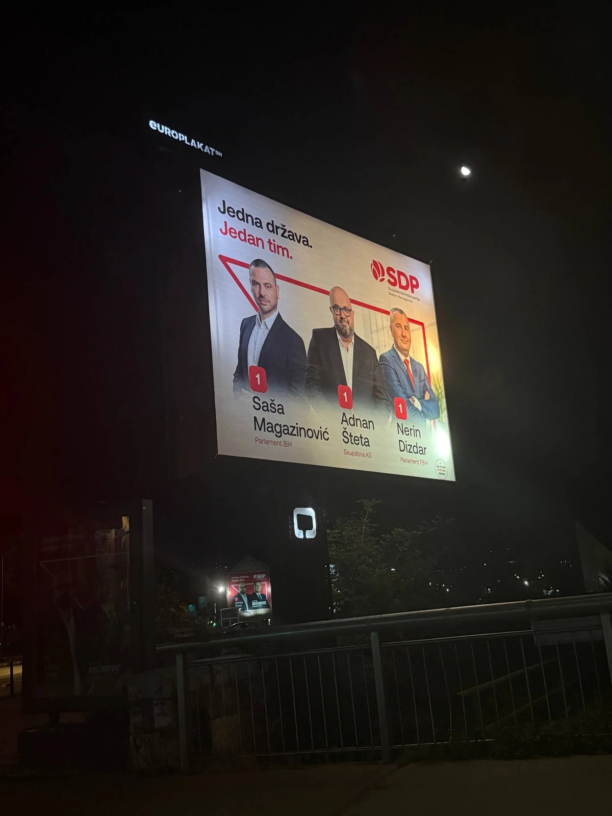 Un grand panneau électoral éclairé la nuit : trois candidats du SDP sous le slogan « Jedna država. Jedan tim. »