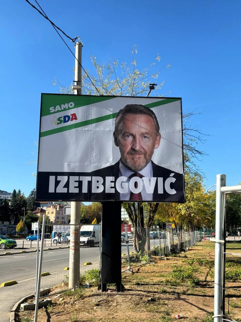 Un panneau électoral du SDA au bord d’une avenue, sous un ciel bleu ; le nom « Izetbegović » en grandes lettres.