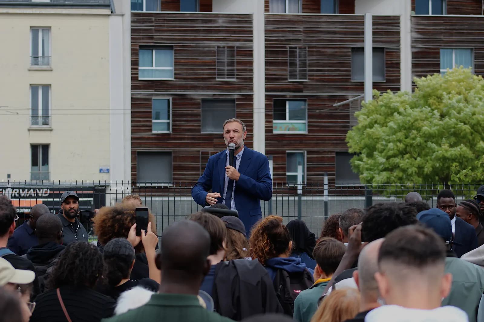 Un homme en costume bleu parle au micro devant une foule rassemblée en plein air, au pied d’immeubles.