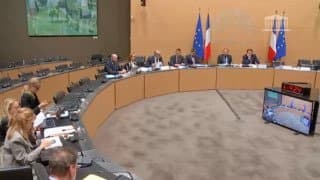 Une salle de commission de l’Assemblée nationale en hémicycle, avec drapeaux français et européens au fond et un écran de retransmission au premier plan.